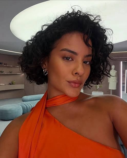 Pessoa posando em um ambiente moderno, vestindo uma blusa laranja com cabelos cacheados. Brinco da Maria de Fátima