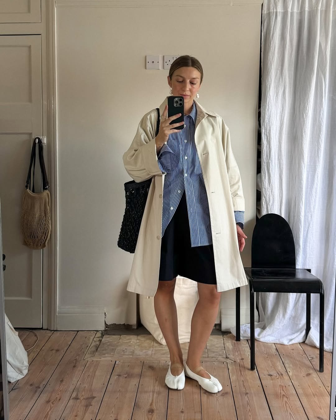 Pessoa com estilo preppy, vestindo camisa listrada azul, casaco claro e sapatos brancos, tirando selfie no espelho.