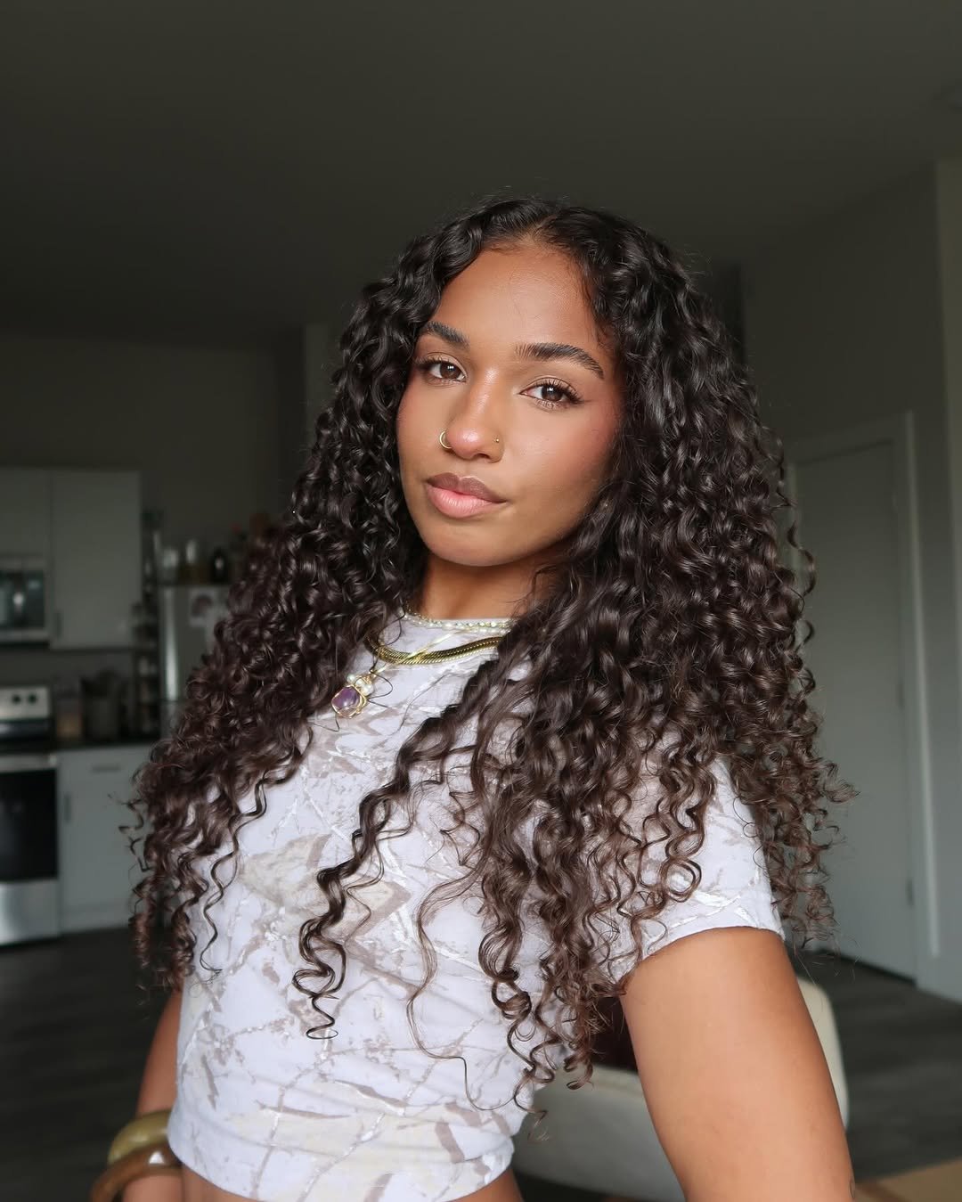 Mulher com cabelo cacheado longo volumoso, usando camiseta clara e colares, em uma sala iluminada.