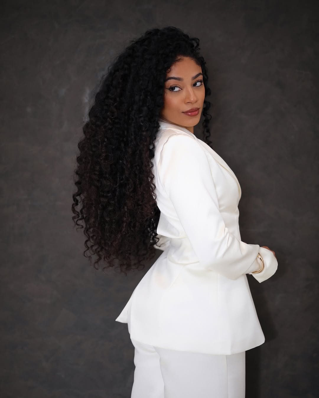 Mulher com cabelo cacheado longo, usando terno branco, olhar voltado para trás em fundo cinza escuro.
