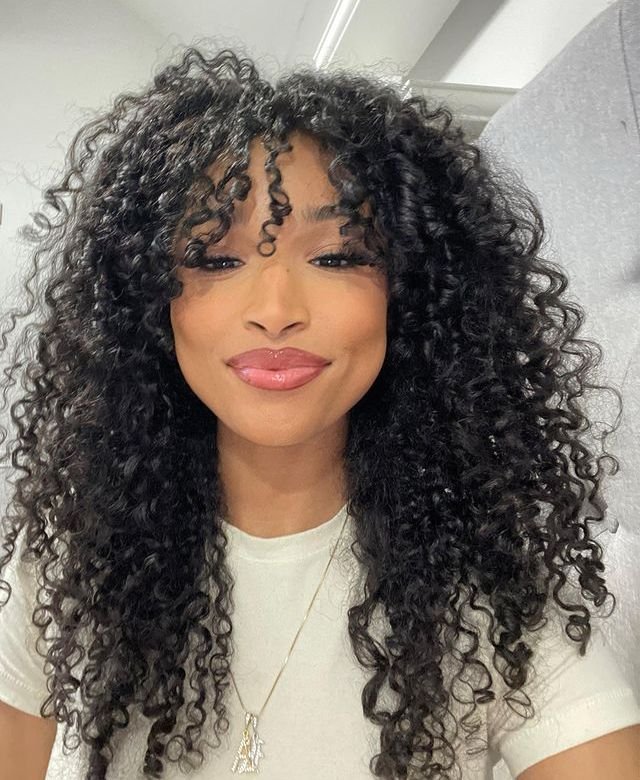 Mulher sorrindo com cabelo cacheado longo, usando blusa clara e colar delicado.