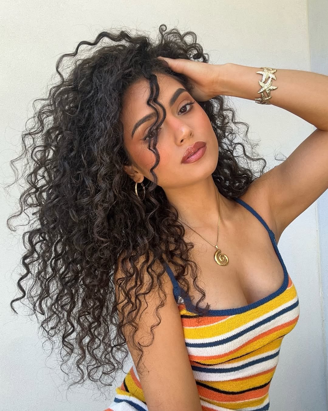 Pessoa com cabelo cacheado longo, usando vestido listrado colorido e acessórios dourados.