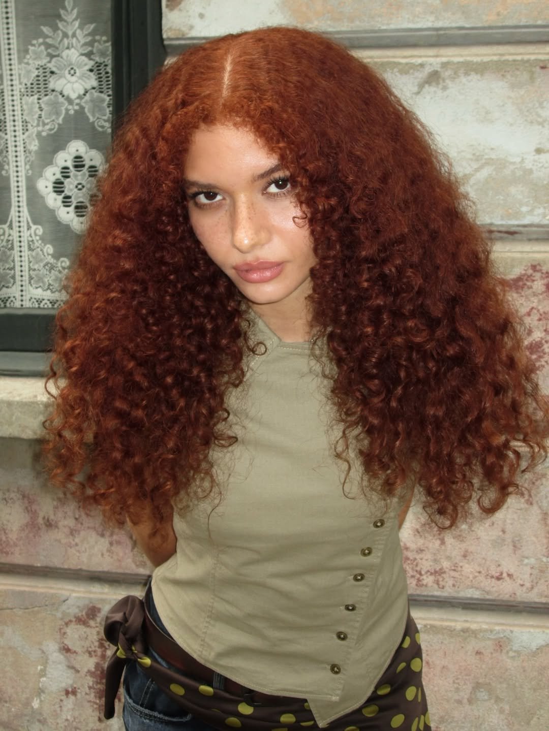 Pessoa com cabelo cacheado longo e ruivo, vestindo camiseta bege e lenço de bolinhas, em frente a uma parede.