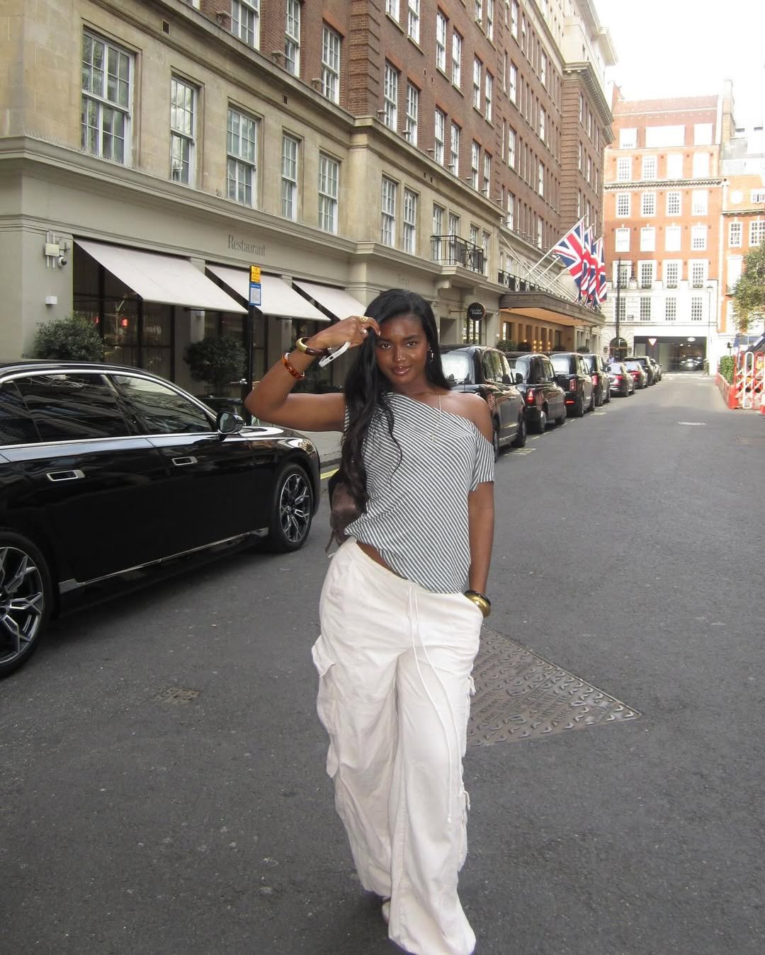 Usuária caminhando em calça branca e camiseta com ombro caído, em rua com prédios e bandeira britânica.