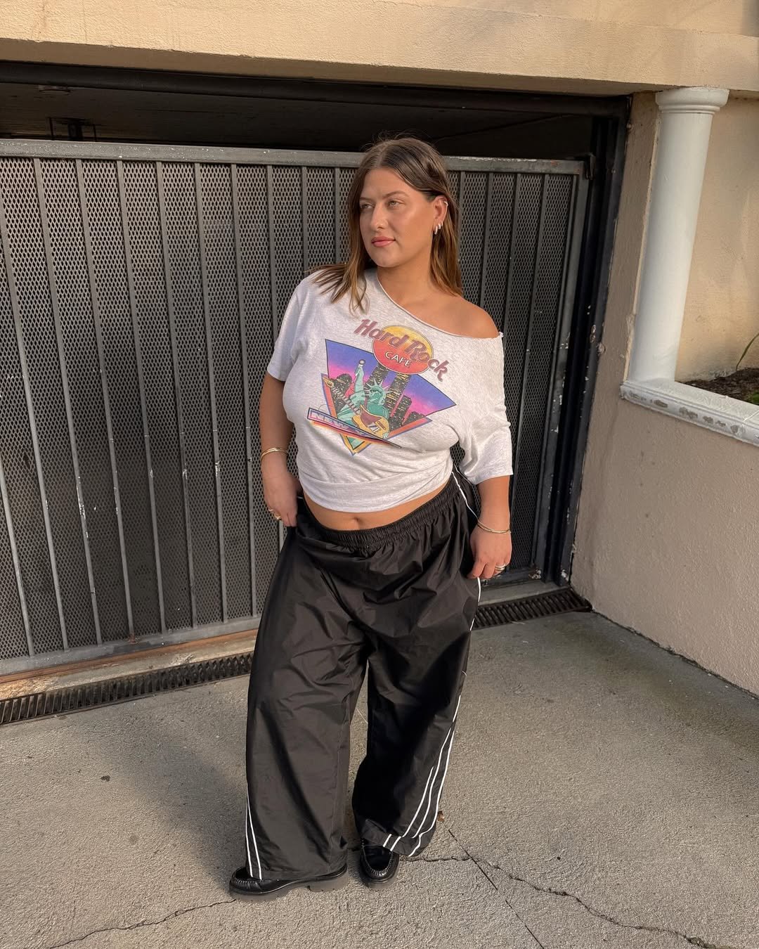 Mulher usando camiseta com ombro caído estampada e calça preta larga em frente a um portão metálico.