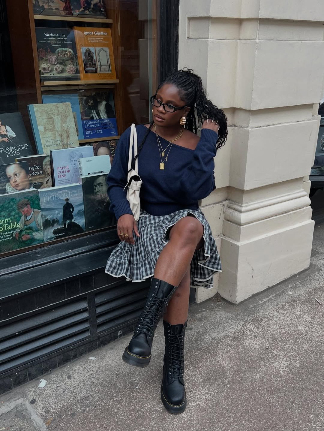 Jovem sentada em frente a uma vitrine, usa camiseta com ombro caído, saia xadrez e botas pretas.