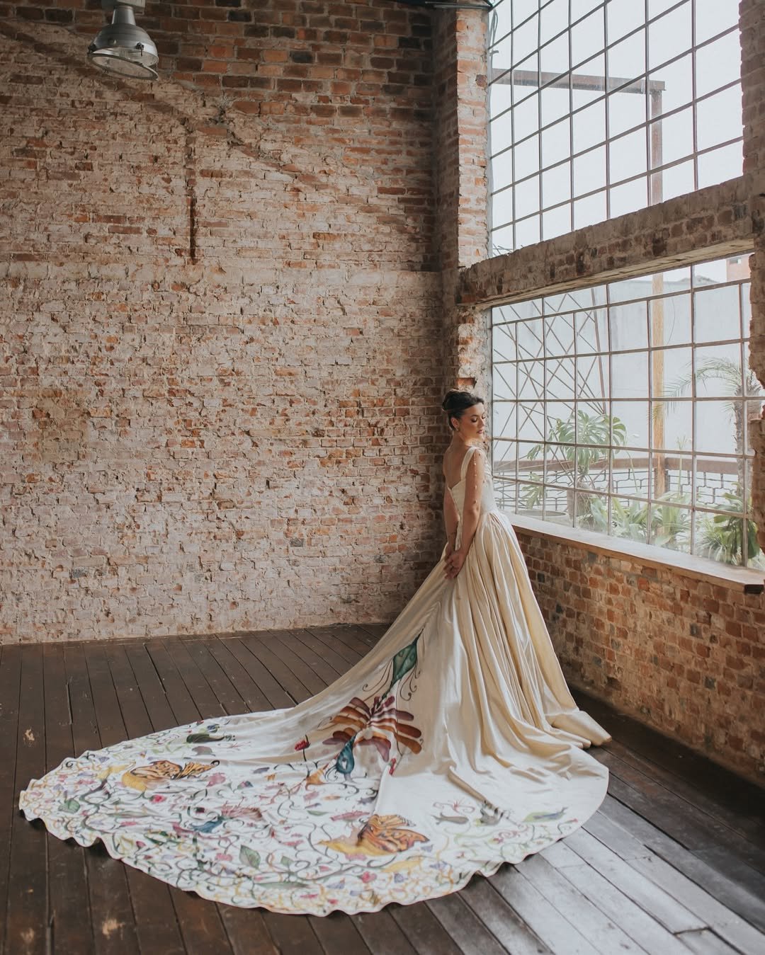 Pessoa em um vestido não tradicional com cauda decorada com estampas coloridas em um espaço rústico com parede de tijolos.