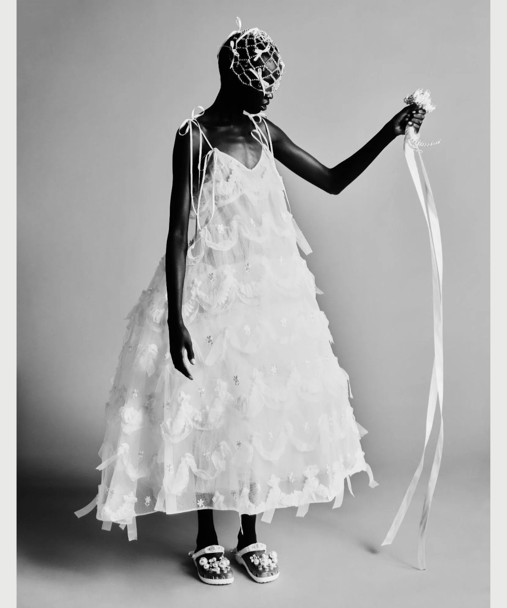 Pessoa vestida em traje não tradicional, com vestido branco volumoso, máscara decorativa e segurando fitas compridas.