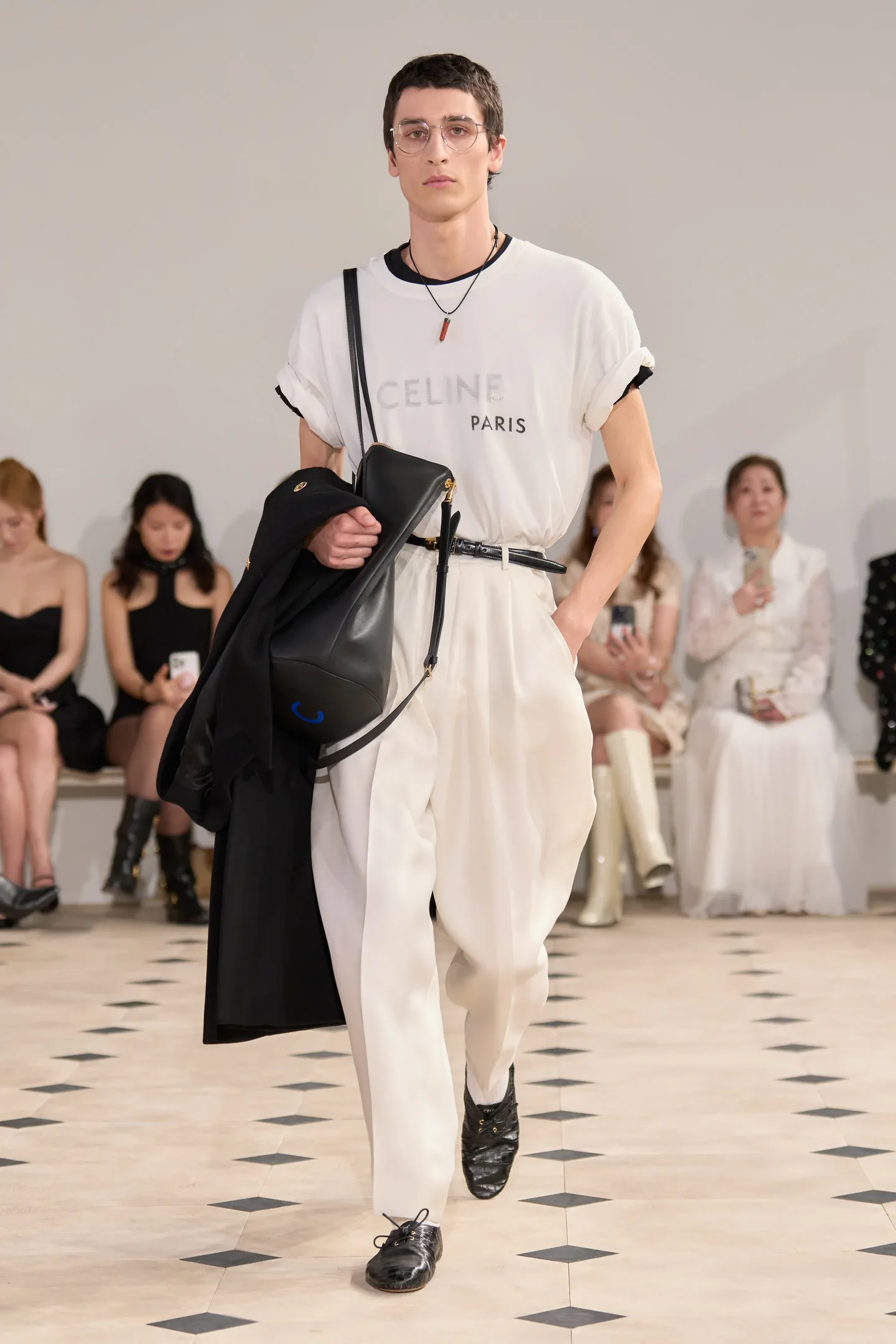 Modelo usando graphic tee com logo, calça branca solta e segurando jaqueta preta em um desfile de moda.