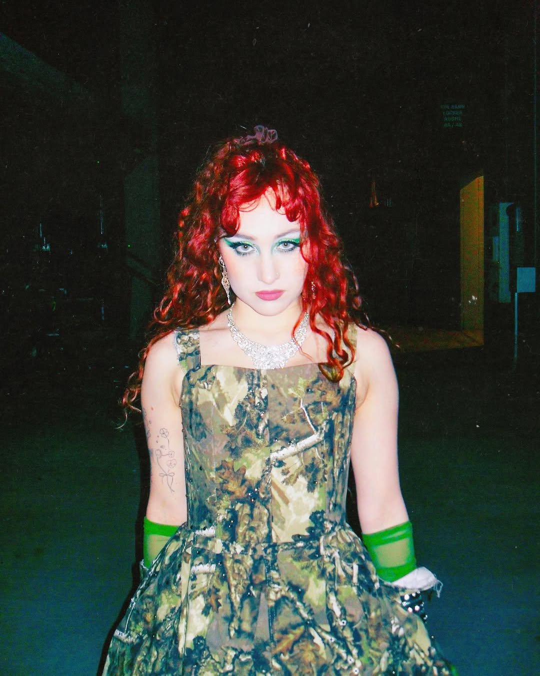 Pessoa com vestido camuflado em ambiente escuro, cabelo vermelho e maquiagem marcante.