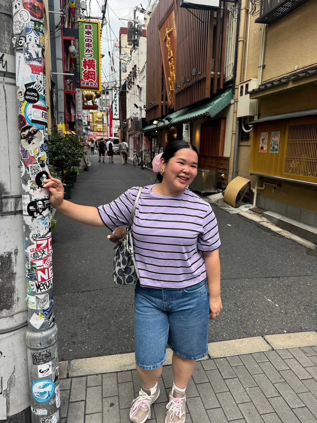 Jovem sorrindo em rua movimentada, usando camiseta listrada, bermuda jeans e chunky sneakers, apoiando-se em poste com adesivos.
