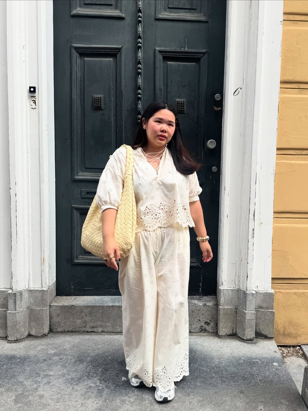 Pessoa em roupa branca elegante, carregando bolsa grande. Usa chunky sneaker, em pé em frente a uma porta escura.