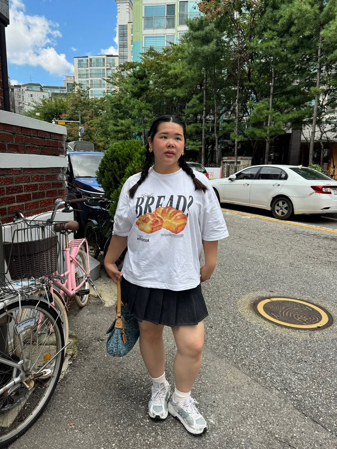 Jovem usando camiseta com estampa de pão, saia preta e chunky sneaker, segura uma bolsa azul em rua arborizada.