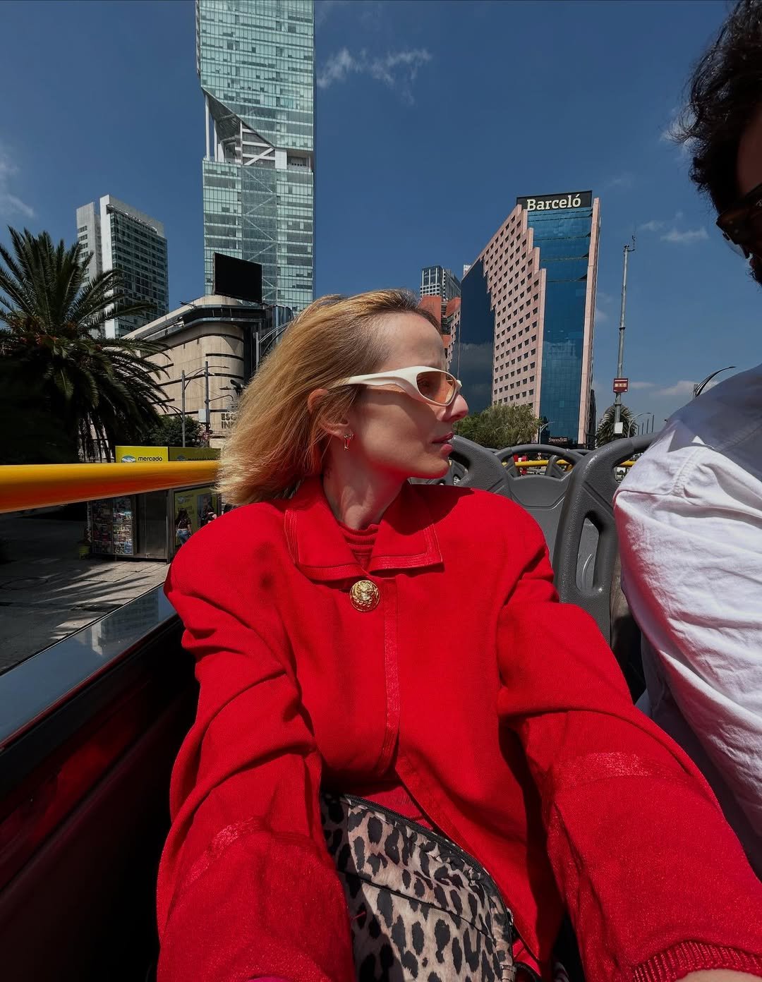 Mulher de casaco vermelho em ônibus turístico, com modernos prédios ao fundo na Cidade do México.