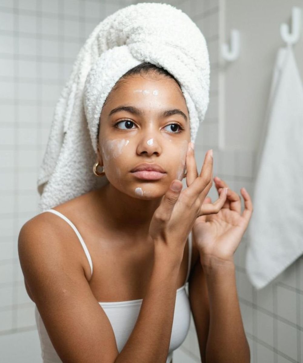 Mulher com toalha na cabeça aplica creme facial no banheiro depois de aprender como acabar com cravos do rosto