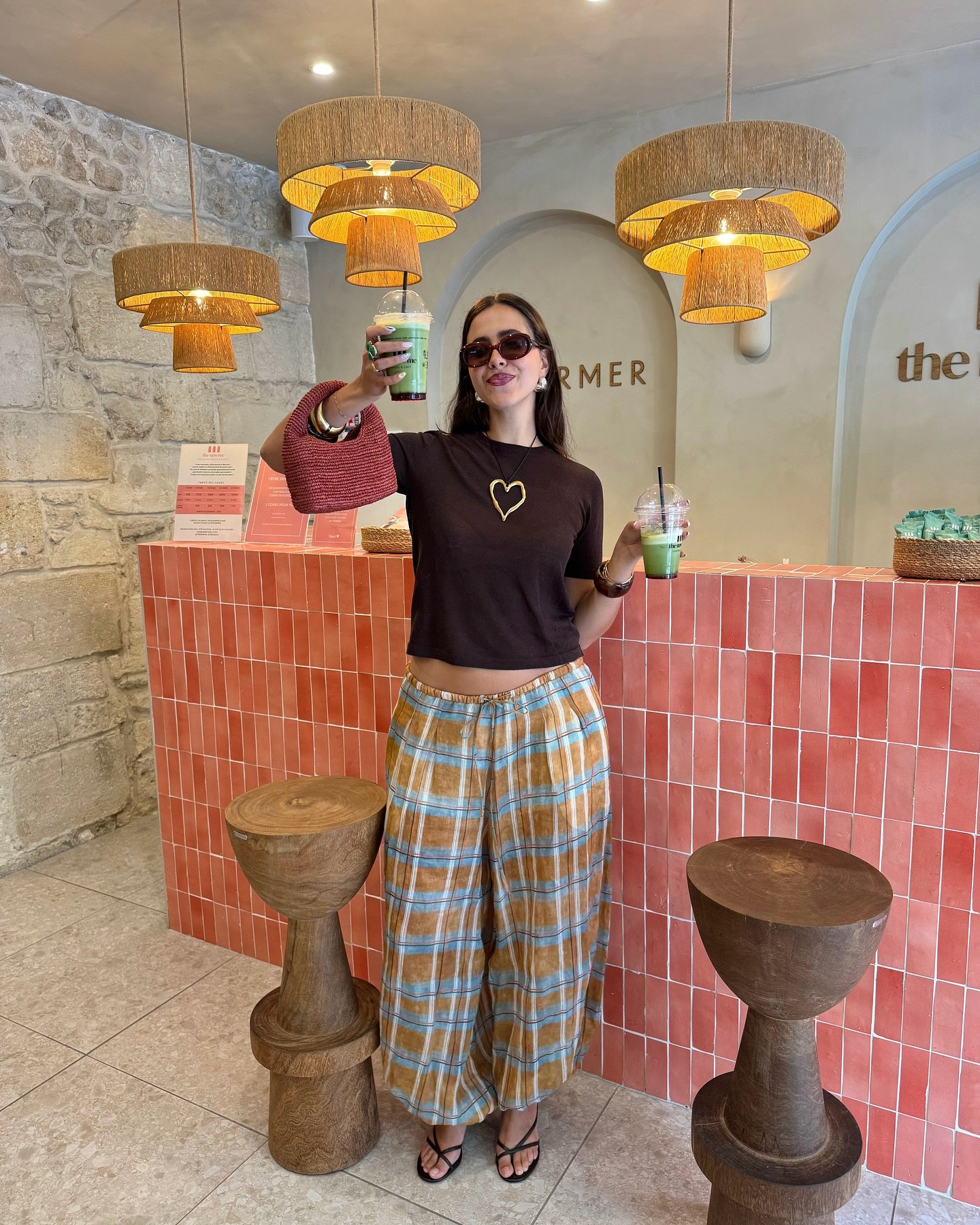 Pessoa vestindo blusa marrom e calça larga xadrez segura bebida em cafeteria com decoração moderna.