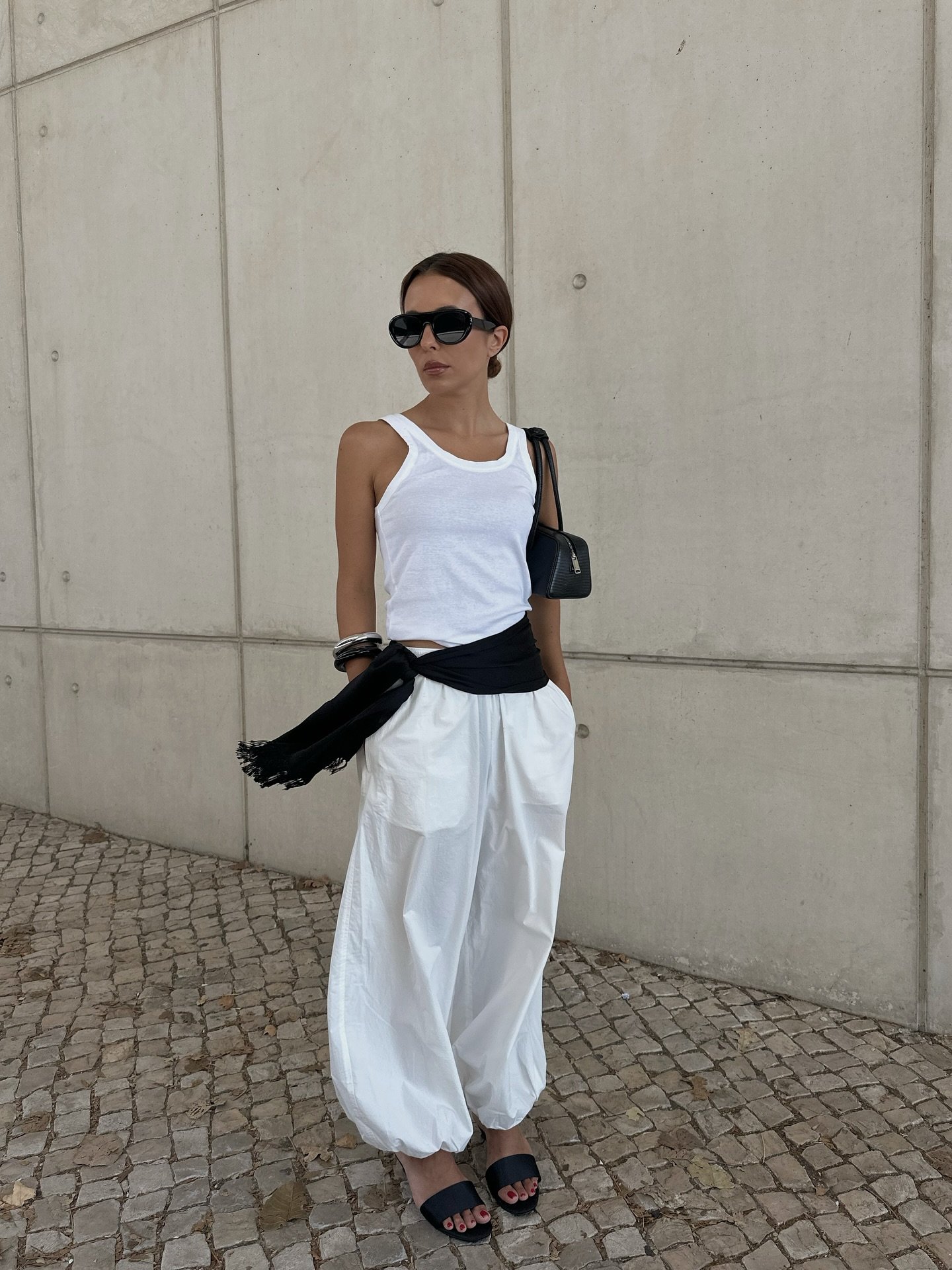Pessoa com blusa branca, óculos escuros e calça aladim branca, posando em frente a uma parede de concreto.