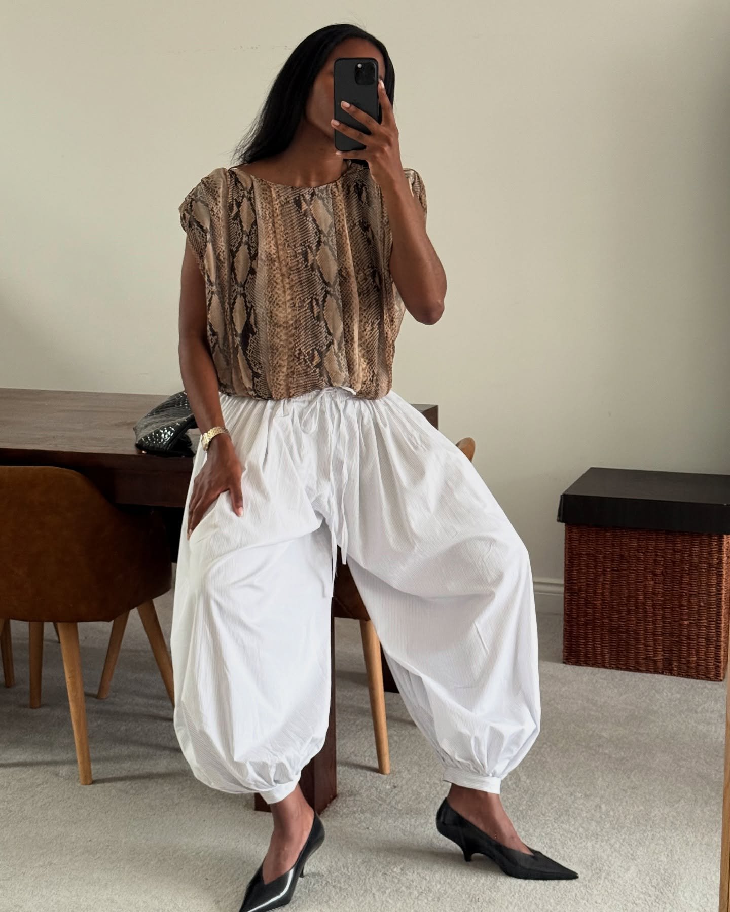 Pessoa com blusa estampada e calça aladim branca, usando sapatos pretos de salto médio, sentada perto de uma mesa.