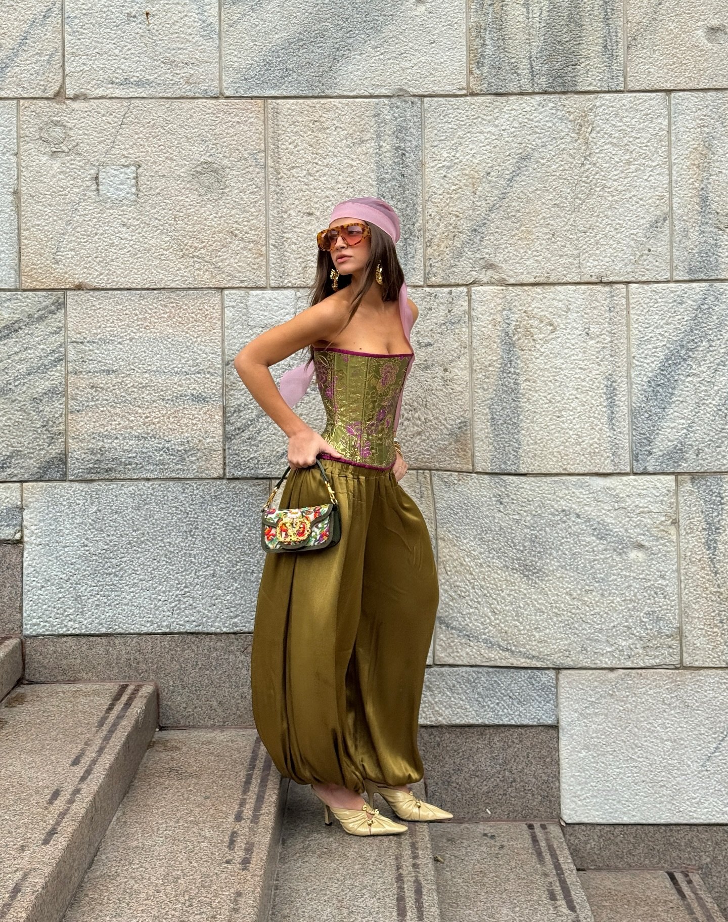 Mulher usando calça aladim verde e corselet estampado. Acessórios incluem óculos, lenço rosa e bolsa colorida.