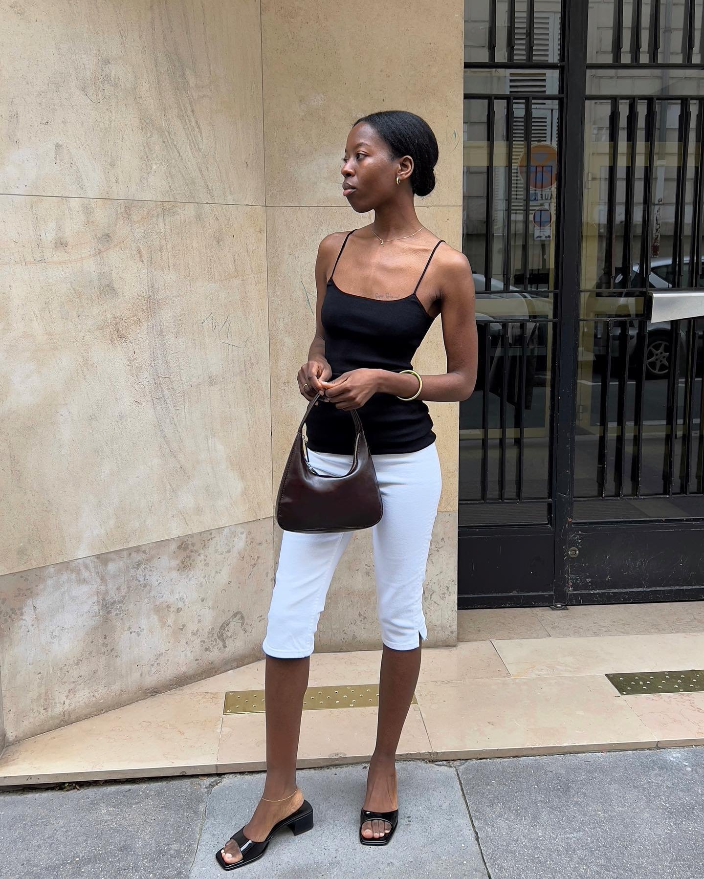 Jovem usando blusa preta e calça corsário branca, segurando bolsa marrom, em frente a um prédio.