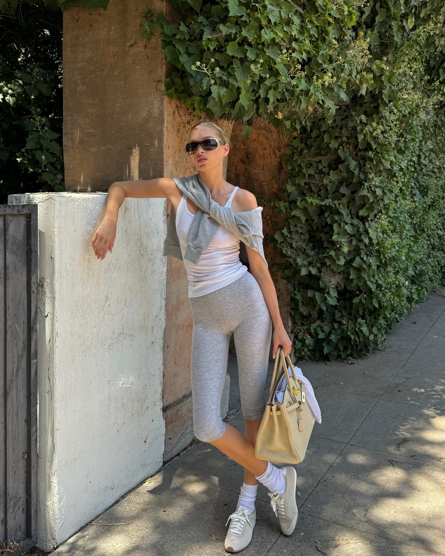 Pessoa usando calça corsário cinza e camiseta branca, encostada em muro com plantas. Segura bolsa bege.