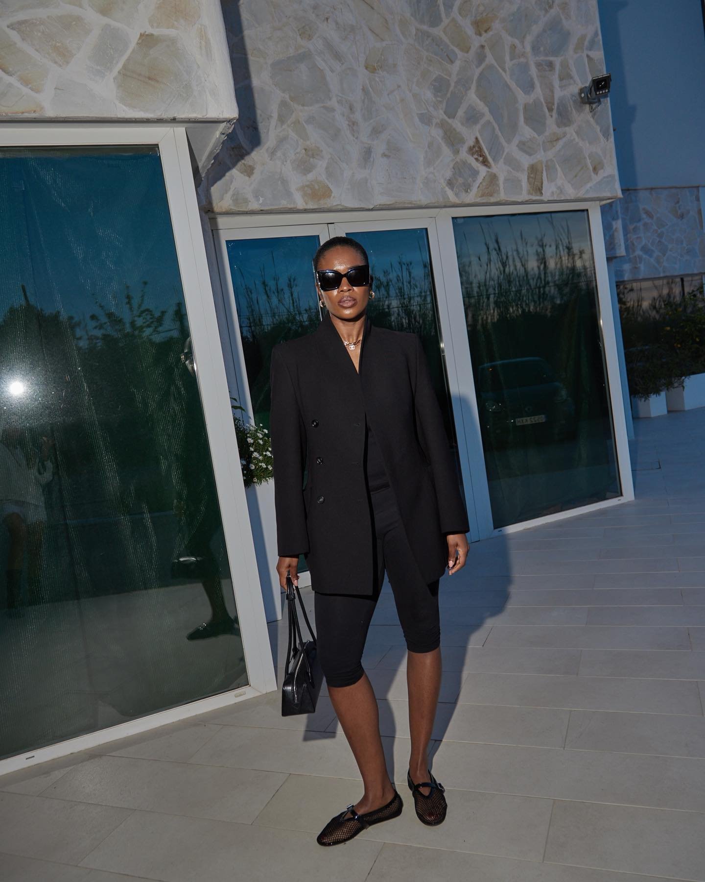 Pessoa de óculos escuros veste blazer preto e calça corsário, segurando bolsa. Ao fundo, parede de pedra e vidro.