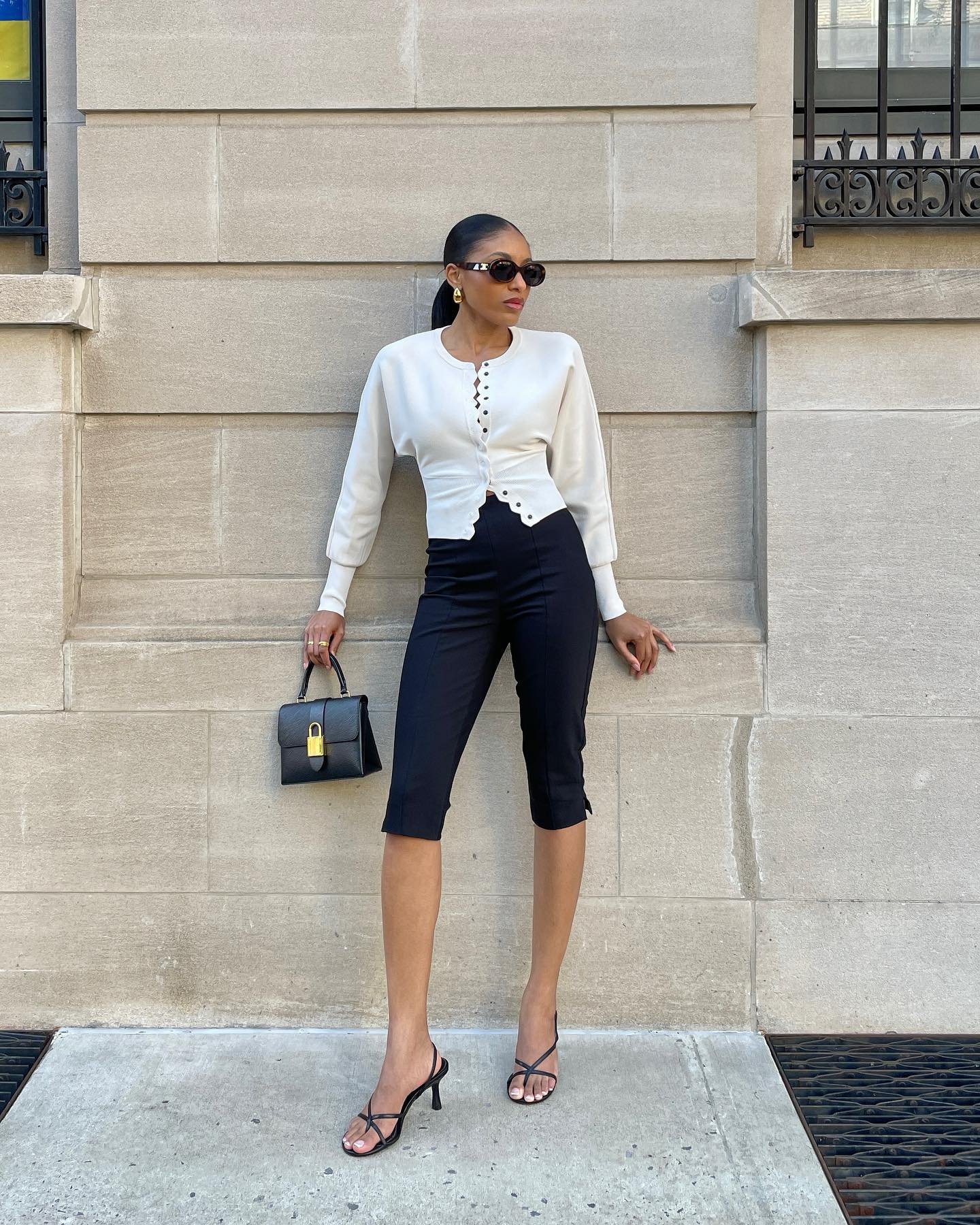 Mulher elegante com casaco branco, calça corsário preta e sandálias de salto, segurando bolsa preta.