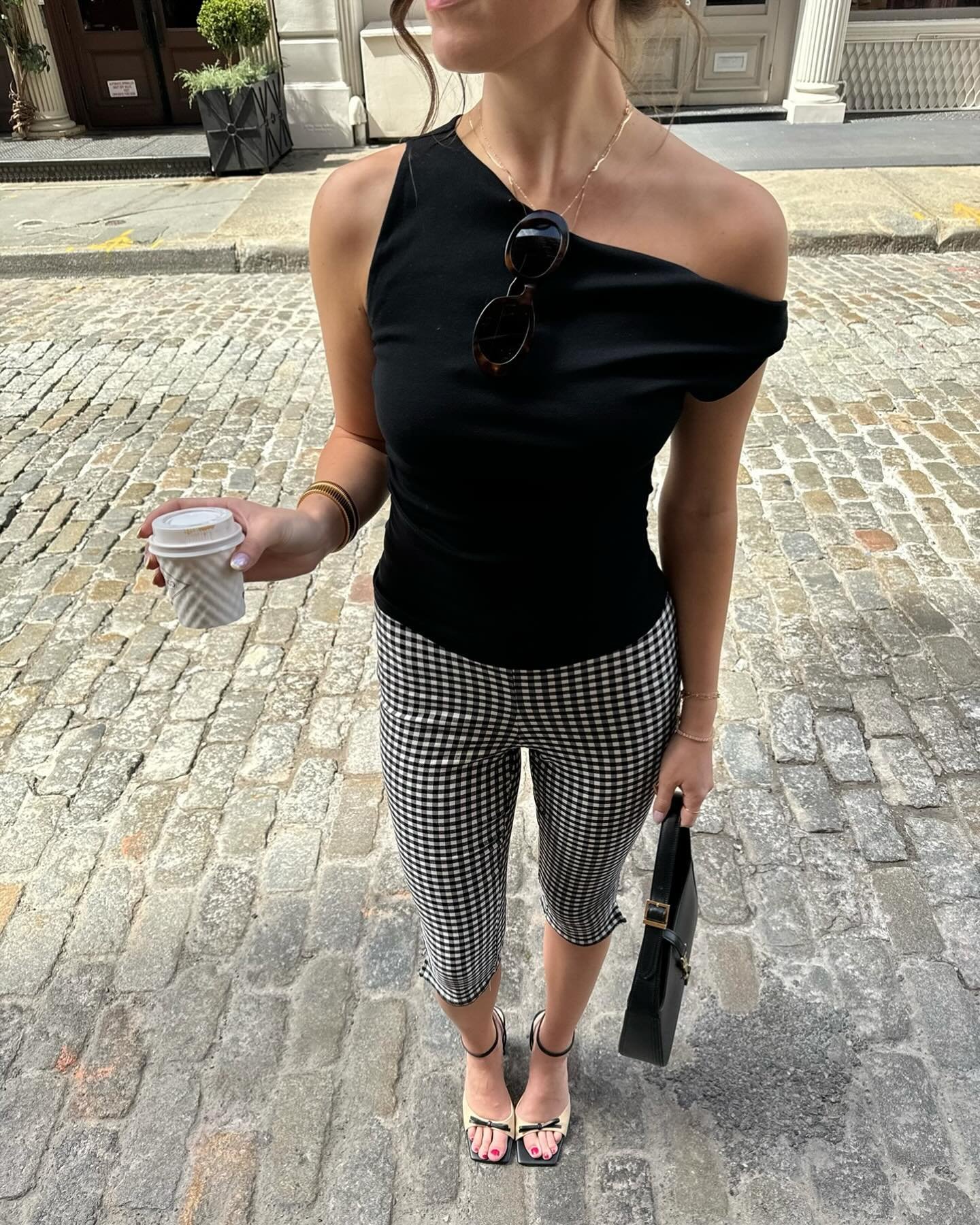 Mulher com blusa preta e calça corsário xadrez, segurando café e bolsa preta, caminha em rua de paralelepípedos.