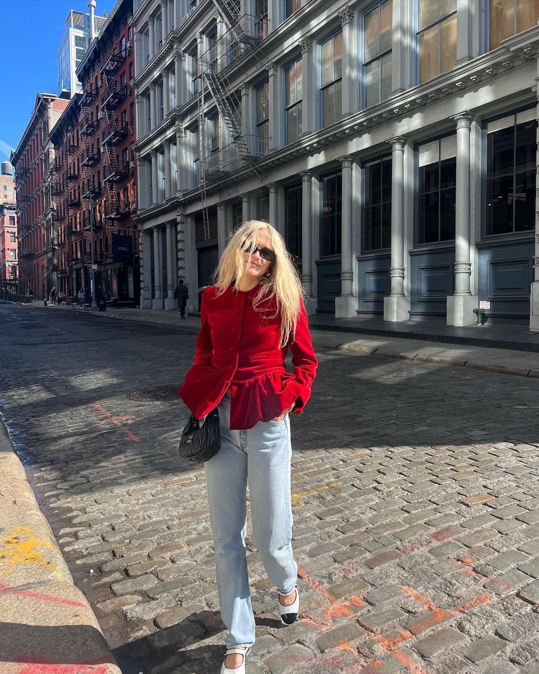 Mulher com blazer vermelho estilo peplum, calça jeans e óculos de sol, andando em rua de paralelepípedos.