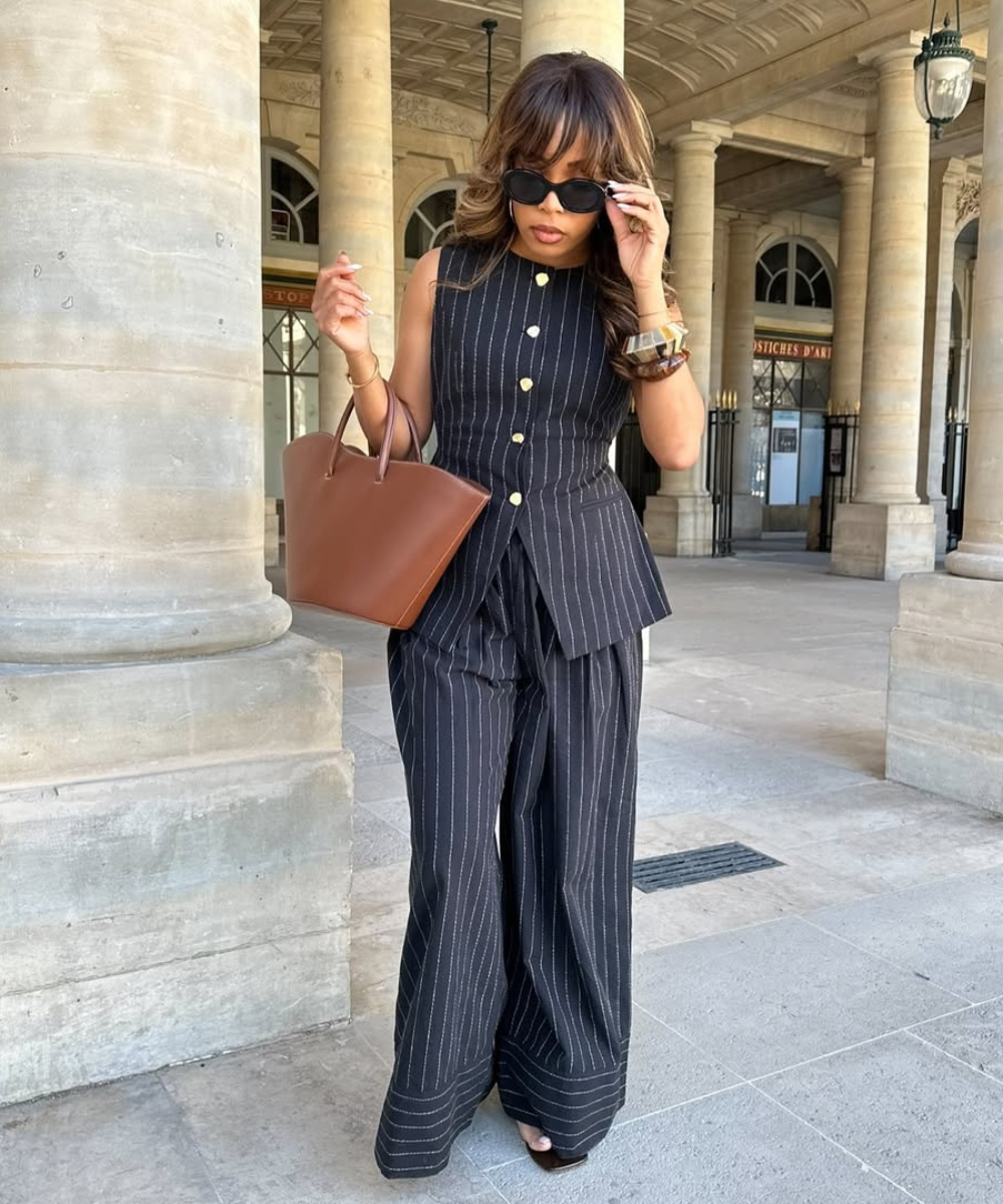 Mulher usando look social sofisticado: conjunto risca de giz, bolsa marrom e óculos escuros, em ambiente externo elegante.