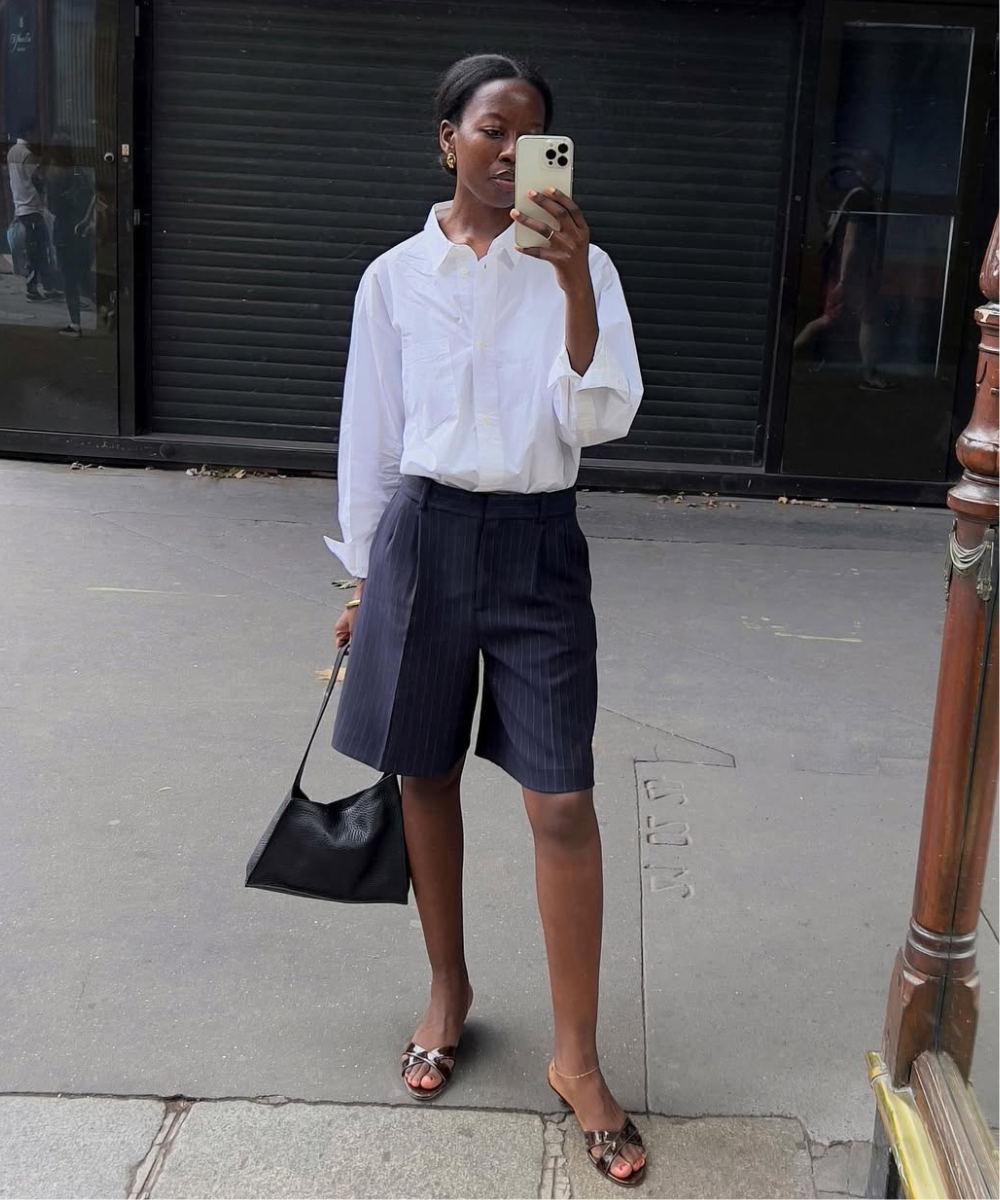 Pessoa em traje social casual com camisa branca e bermuda escura, segurando um smartphone e bolsa preta.