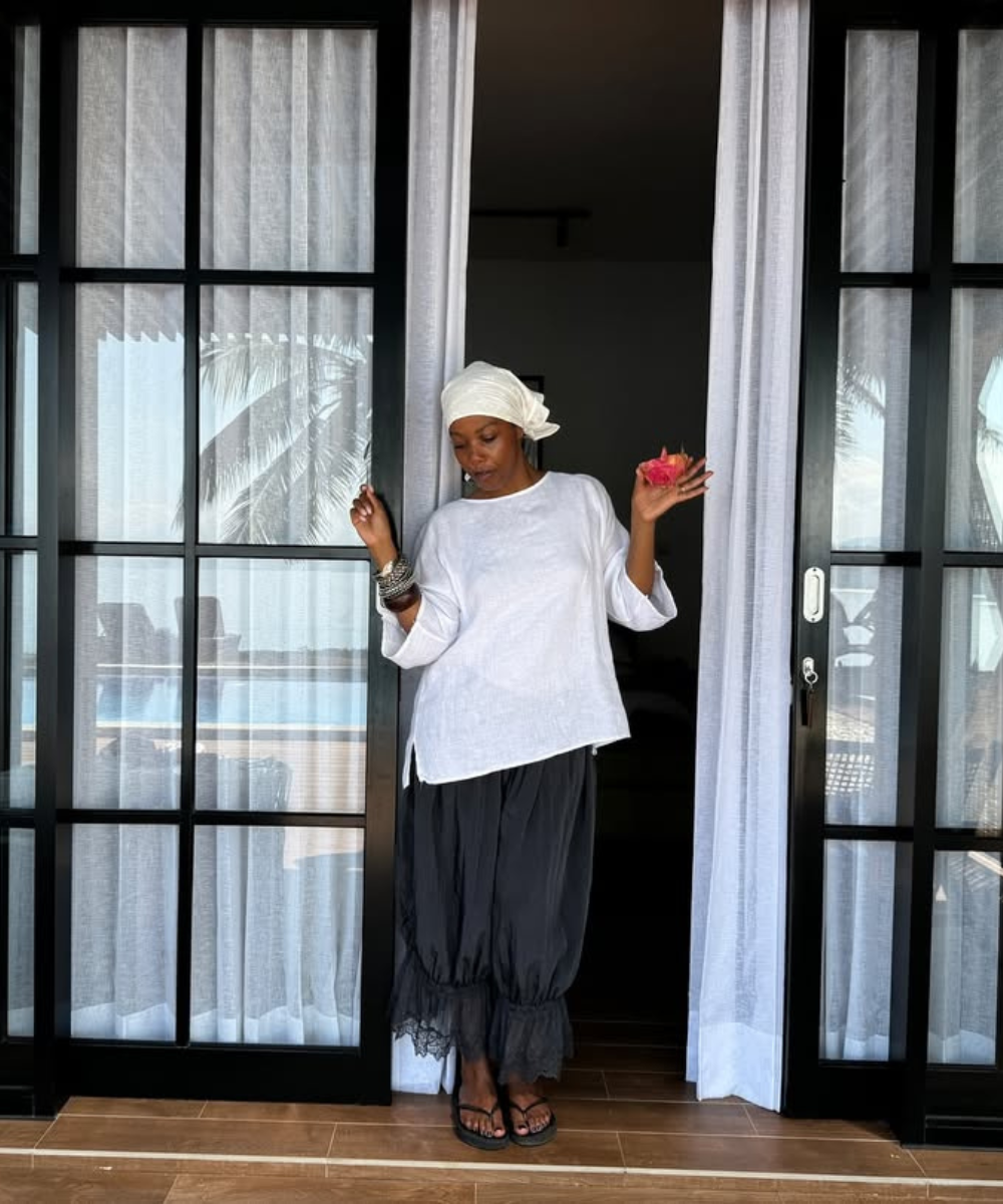 Pessoa com chapéu branco, camiseta solta e saia com detalhe de renda, em um ambiente tropical.