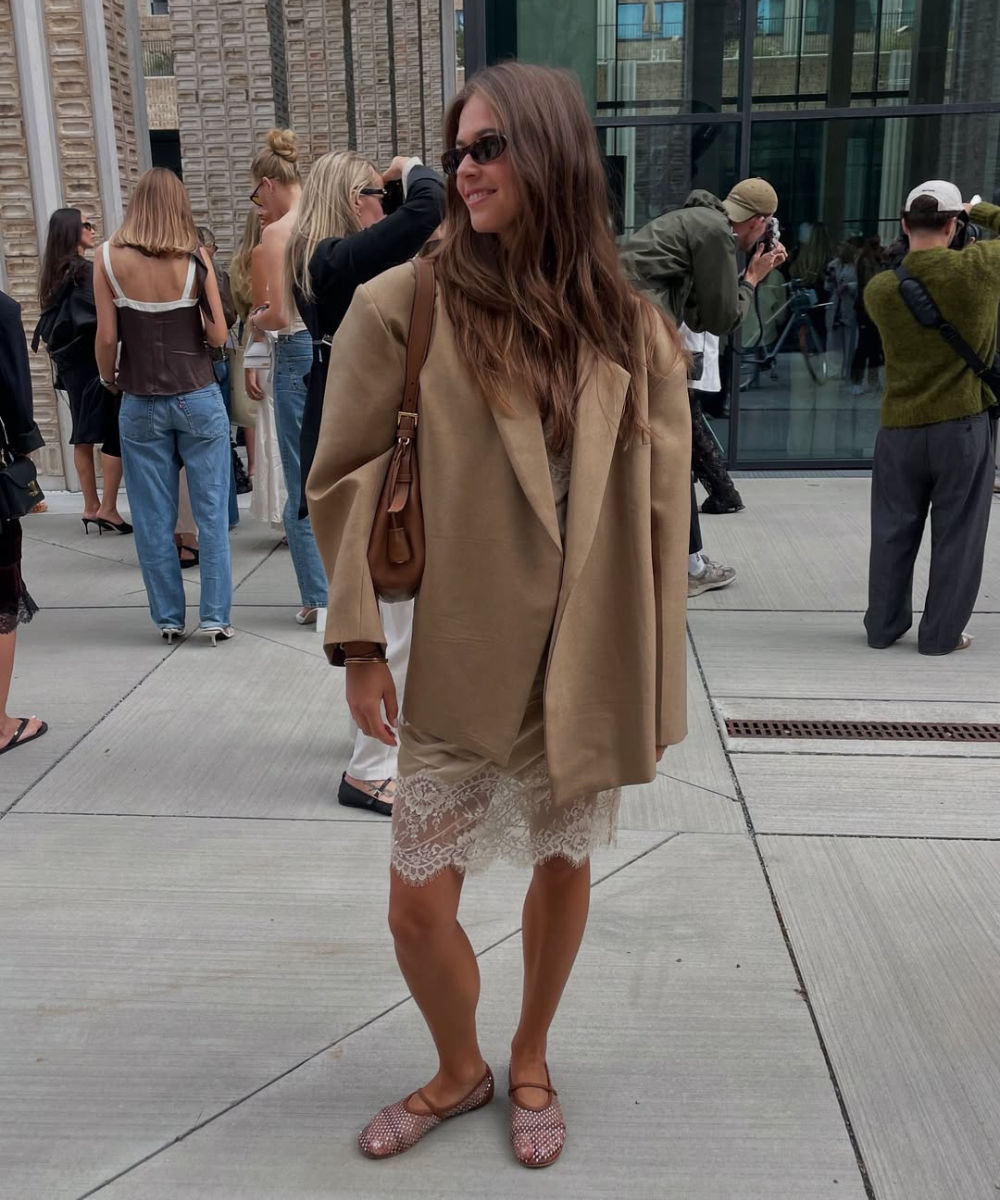 Mulher usando vestido bege com detalhe em renda sob um blazer largo, combinando com sapatilhas marrons.