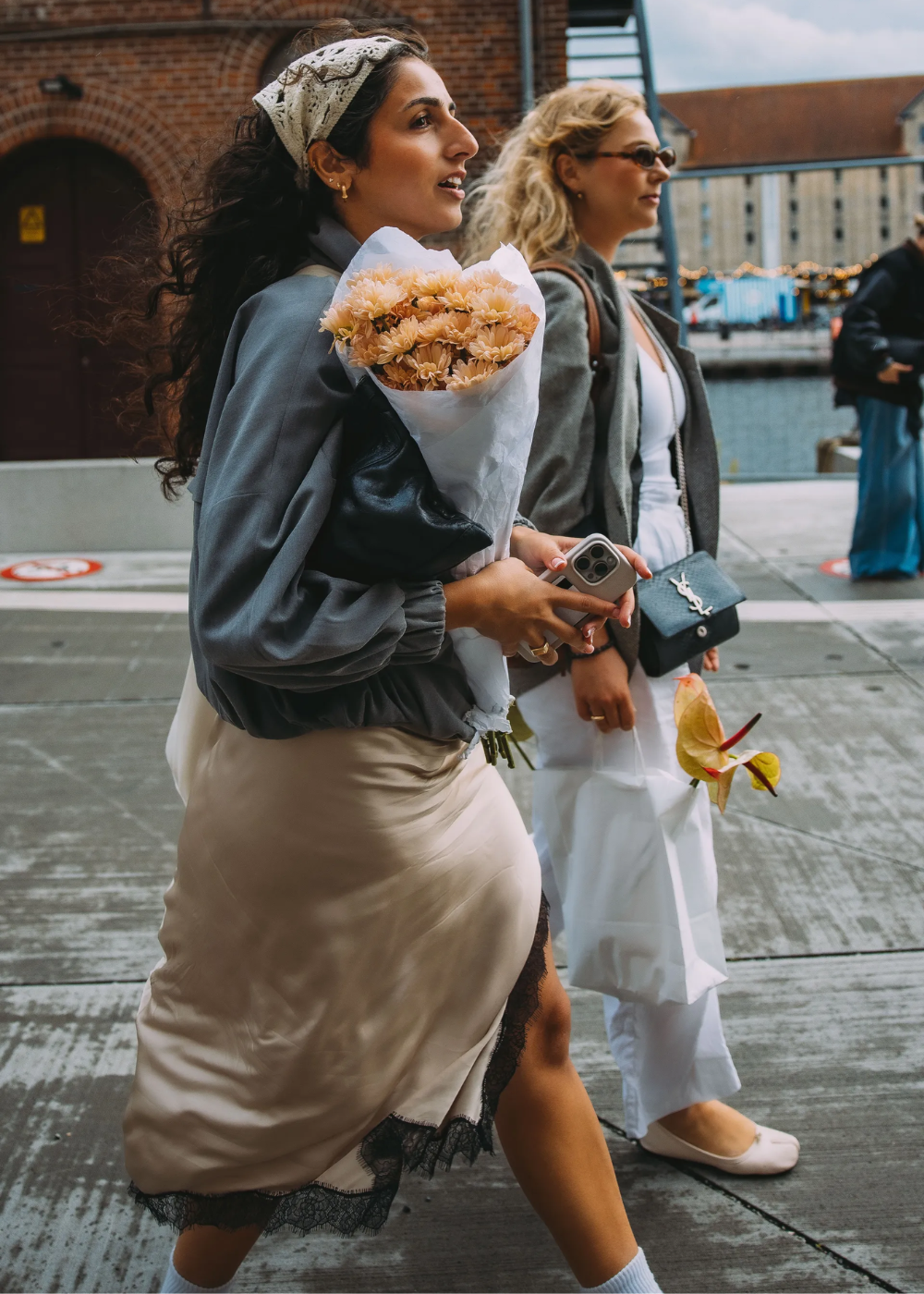 Duas mulheres caminhando; uma com lenço de renda na cabeça e buquê de flores, exibindo estilo e elegância.