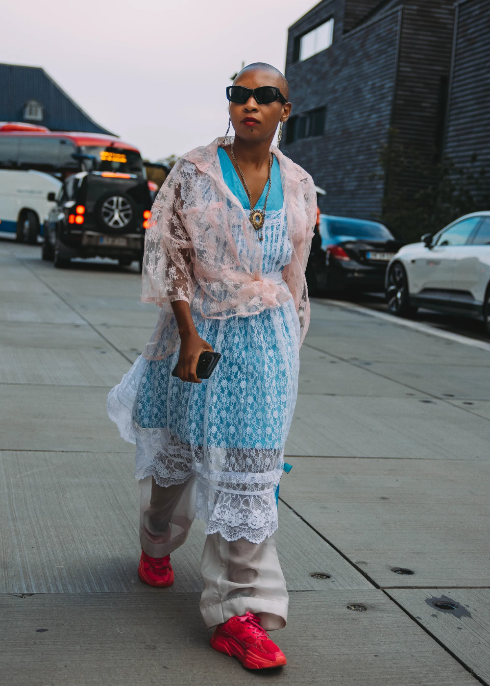Pessoa usando vestido de renda azul sobre calças largas, com tênis rosa vivos e óculos escuros.
