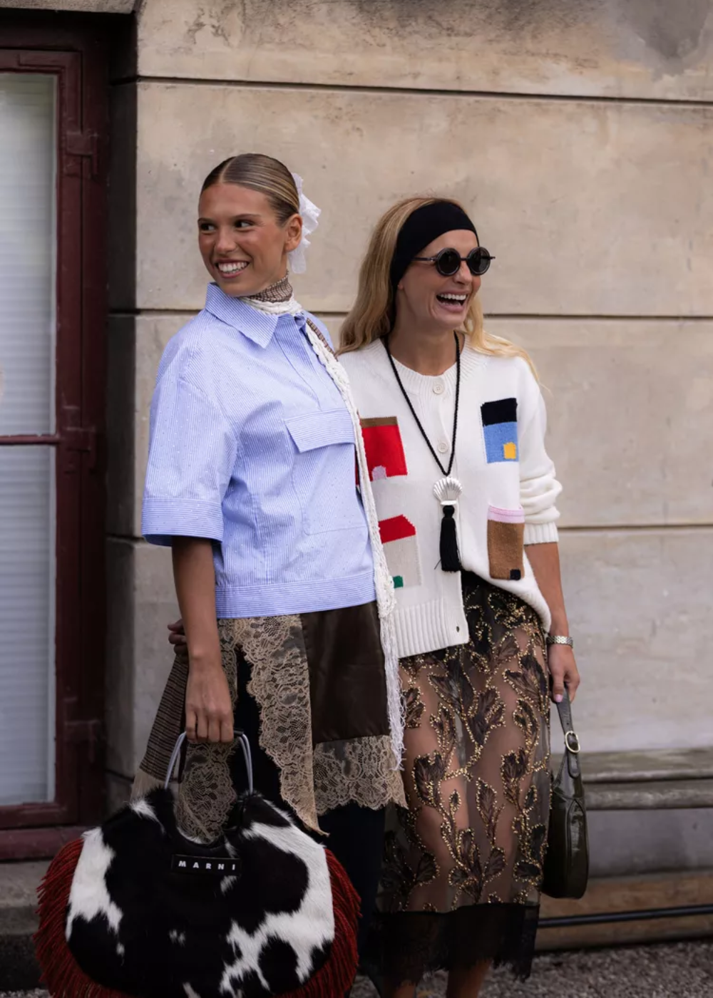 Duas mulheres usam looks que incorporam renda. Uma veste blusa azul com detalhes em renda e bolsa, a outra, saia de renda preta.