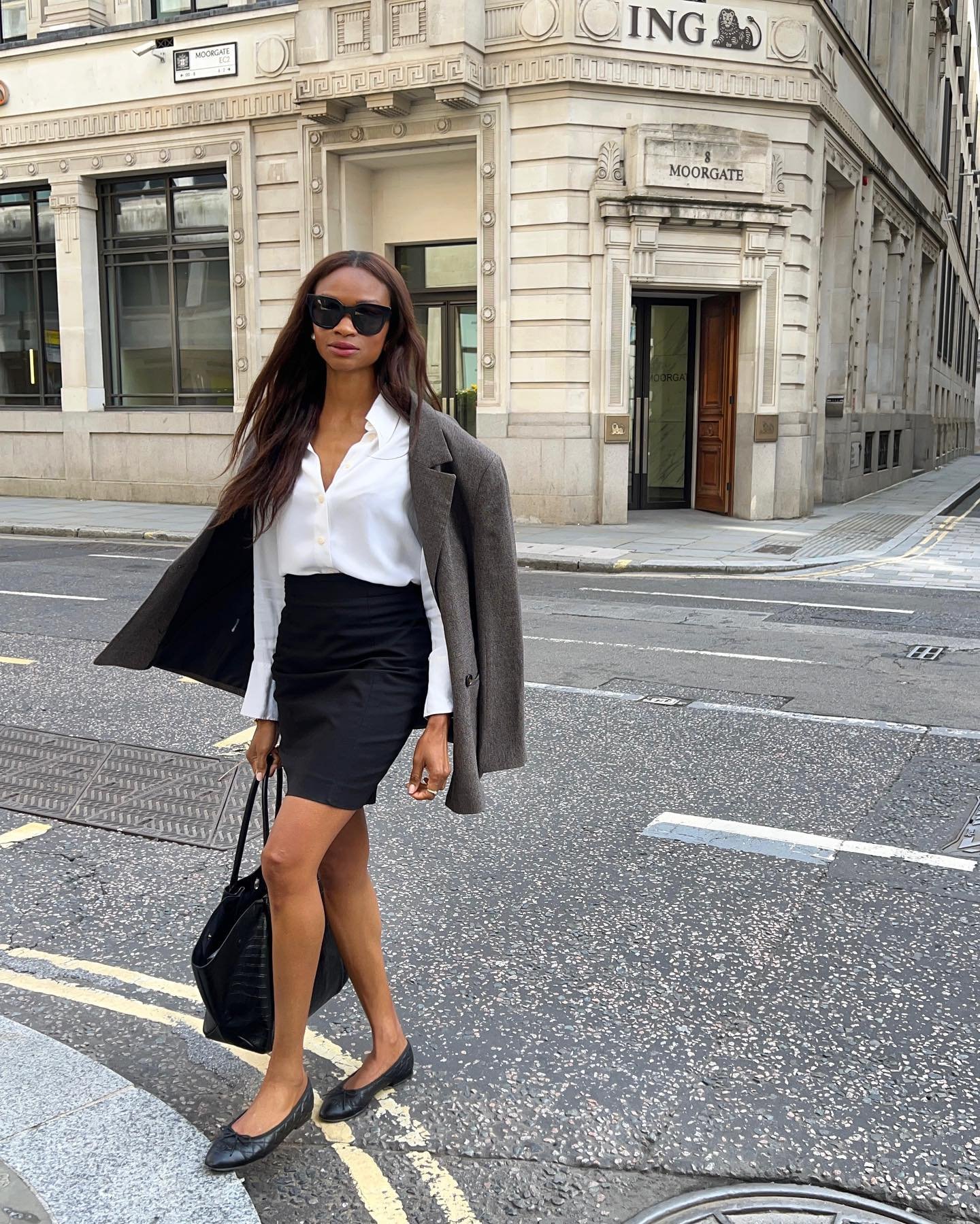 Mulher usando saia para trabalhar preta e camisa branca, com blazer cinza e bolsa, caminha em rua urbana.