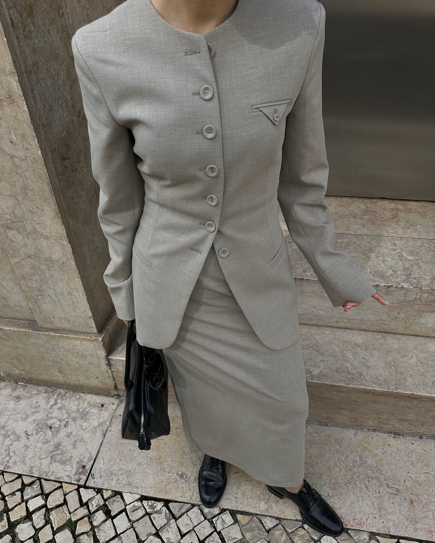 Traje formal feminino cinza com saia longa e blazer de botões. Sapatos pretos e bolsa nas mãos.