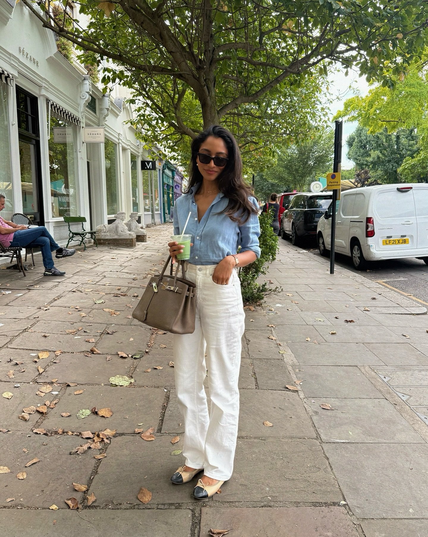 Mulher em rua arborizada com calça branca, camisa azul e sapato com laço, segurando bebida verde.