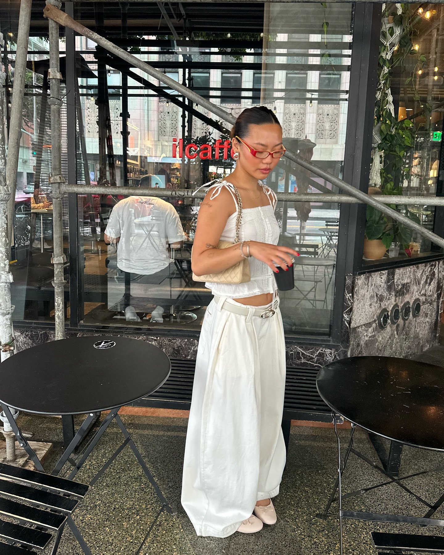 Mulher de óculos vermelhos e roupa branca segura um copo próximo a mesas de café na calçada.
