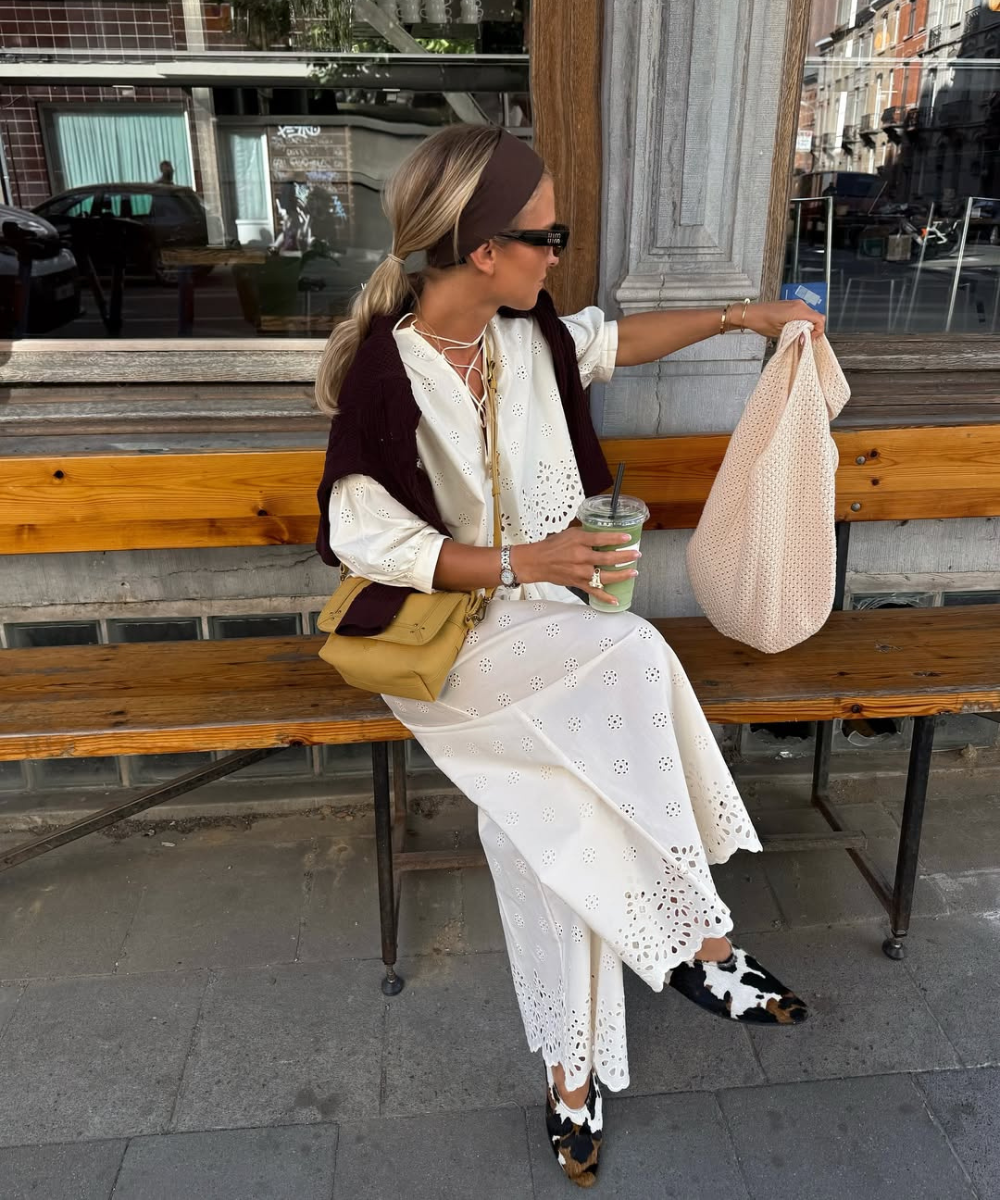 Mulher com vestido branco vazado, bolsa amarela e outra beige, segura bebida verde, com sapatos estampados.