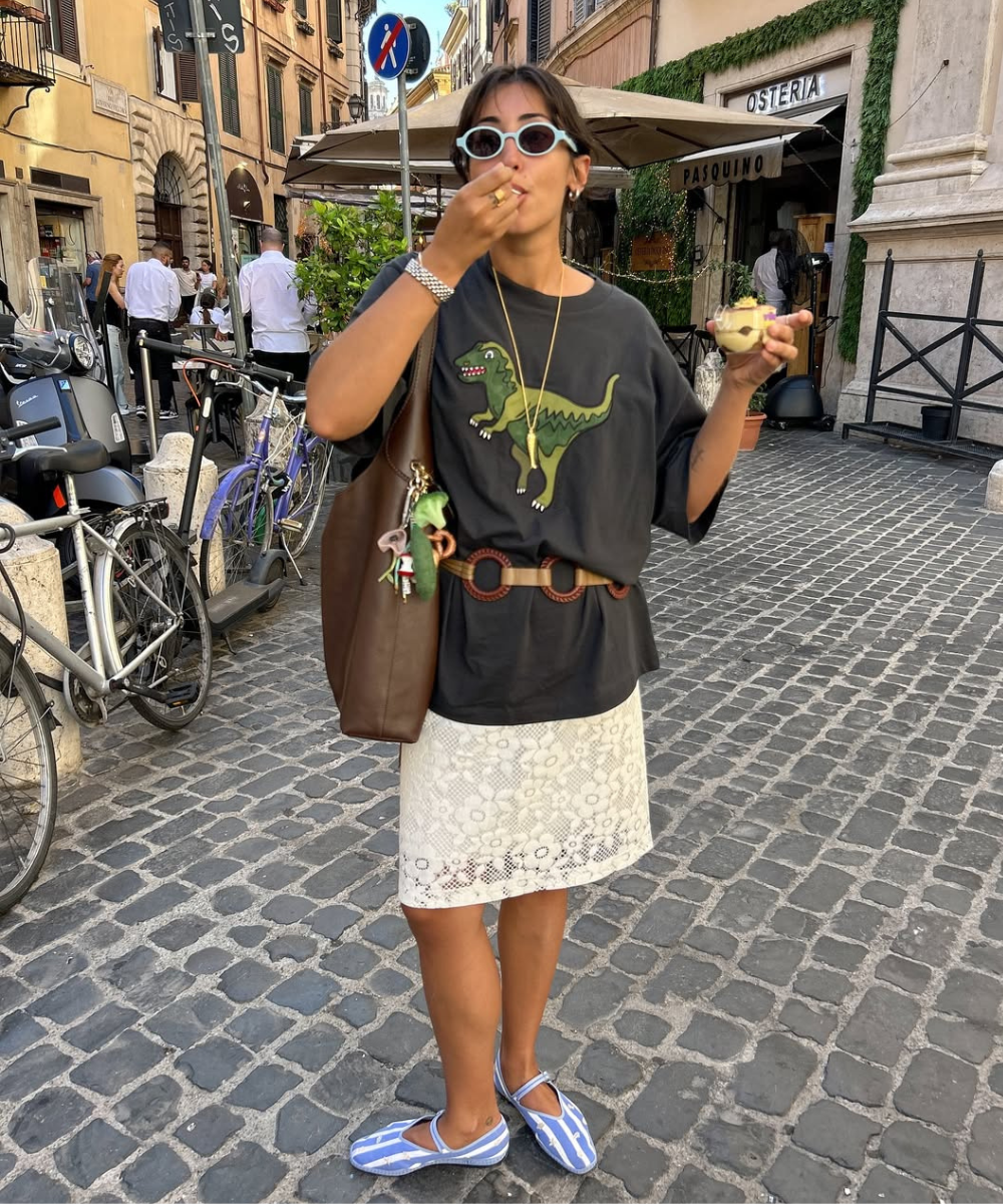 Pessoa usa óculos de sol, camiseta com dinossauro, saia de renda e sapatos azuis. Bolsa marrom, possível tendência entre as cores de bolsa do verão.