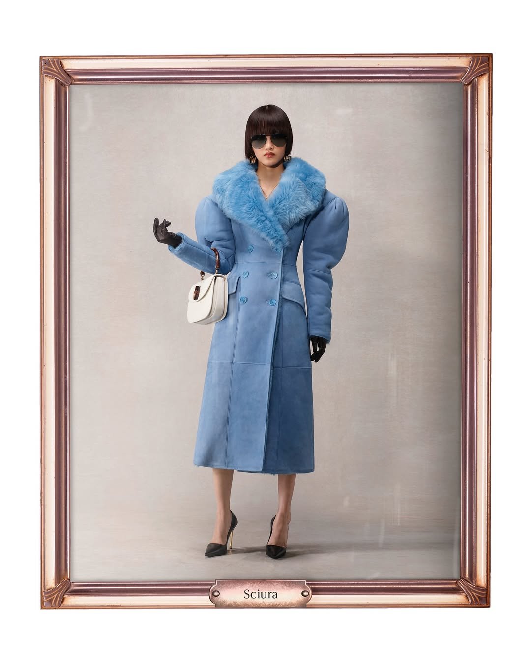 Pessoa vestindo um casaco azul com gola de pele, óculos escuros e luvas, segurando uma bolsa branca. Gucci de Demna