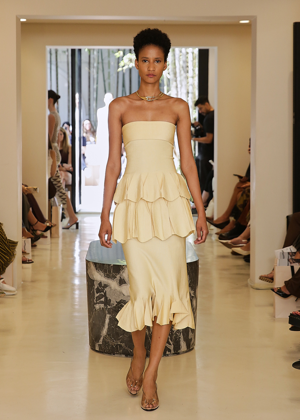 Modelo com vestido bege de babados desfilando em evento de moda LOLITTA em espaço elegante.