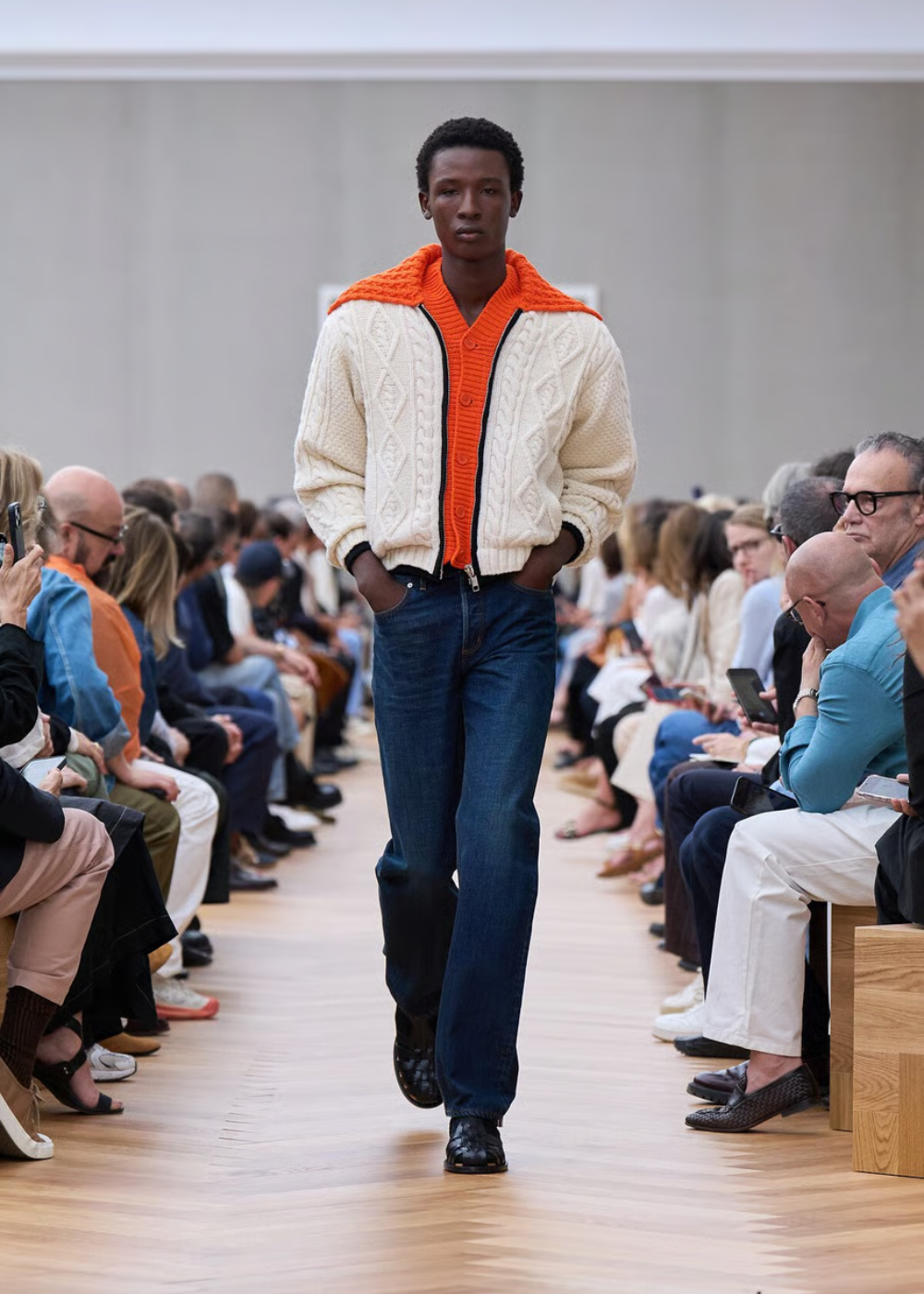 Modelo desfila usando suéter trançado creme e blusa laranja, estilo casual e preppy, em passarela cercada por espectadores.