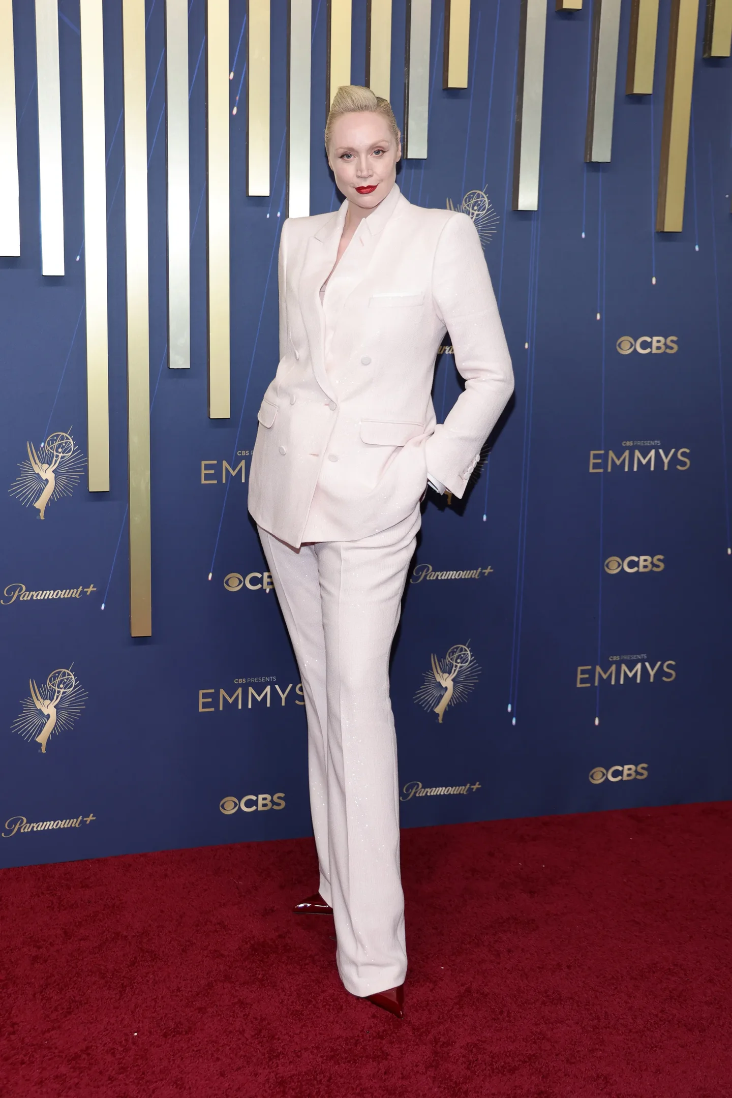 Pessoa em terno elegante branco no tapete vermelho do Emmy 2025, com fundo azul e logotipo do evento.