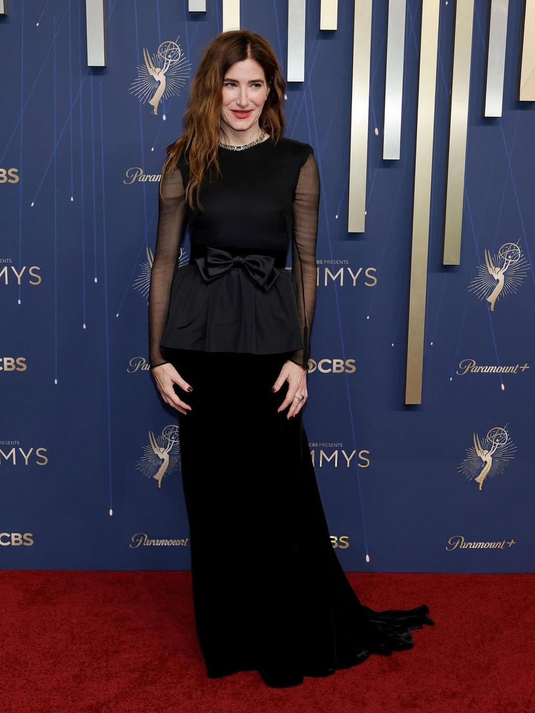 Mulher em vestido preto no tapete vermelho do Emmy 2025, com fundo decorado com logotipos do evento.