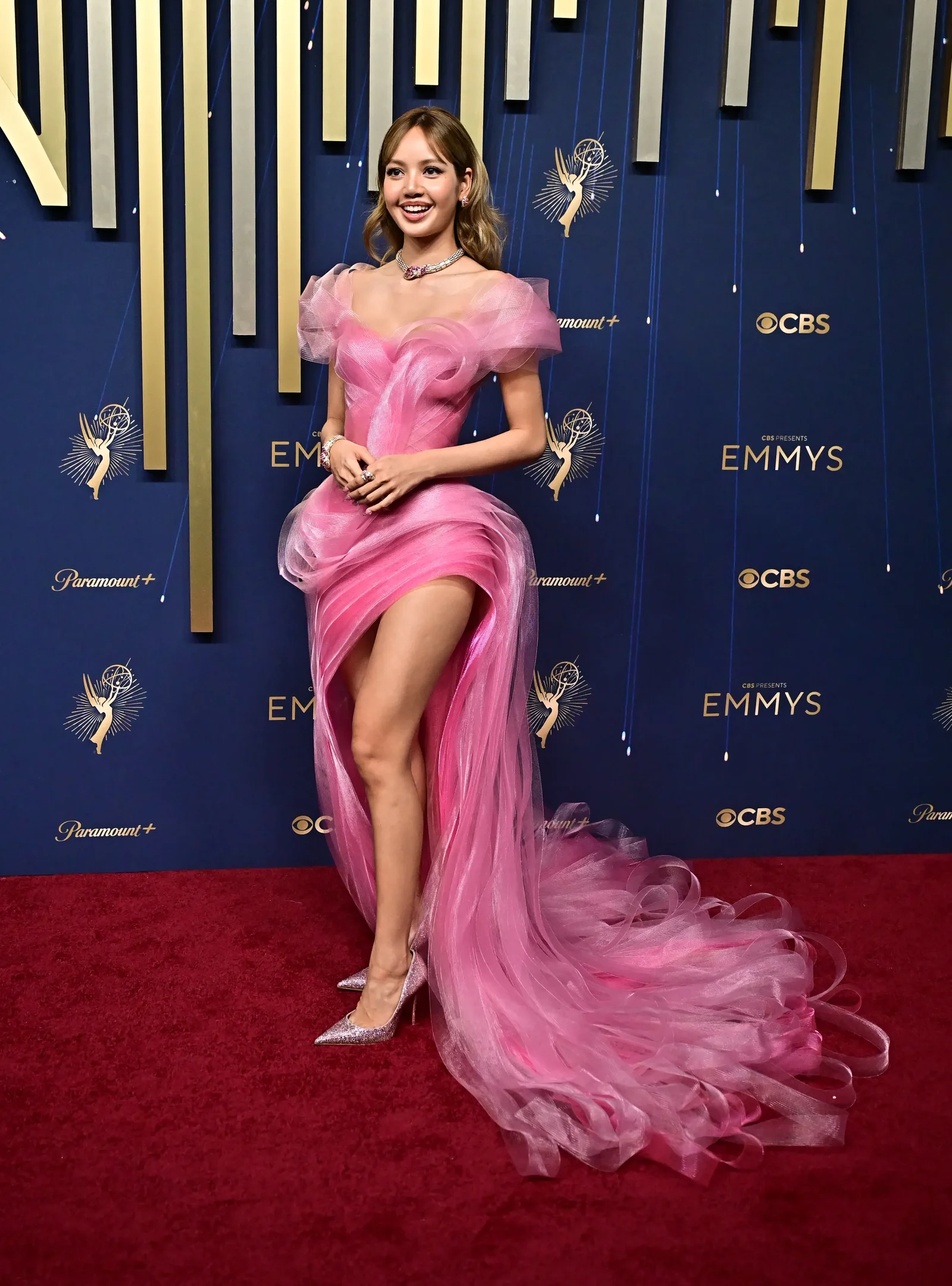 Celebridade usa vestido rosa elegante no evento Emmy 2025, com decoração dourada ao fundo.