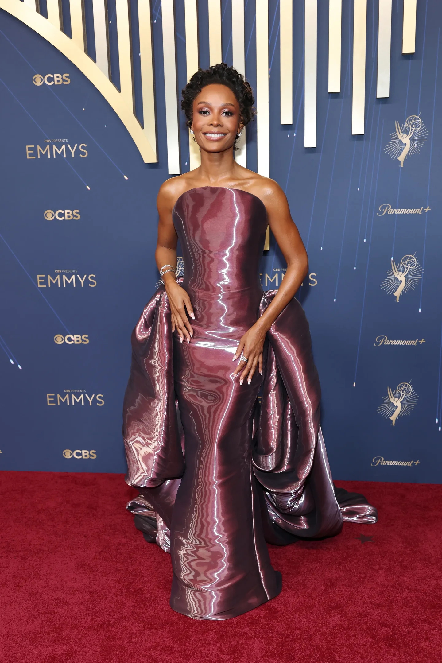Celebridade usando vestido brilhante na cerimônia do Emmy 2025, em tapete vermelho com fundo decorativo.