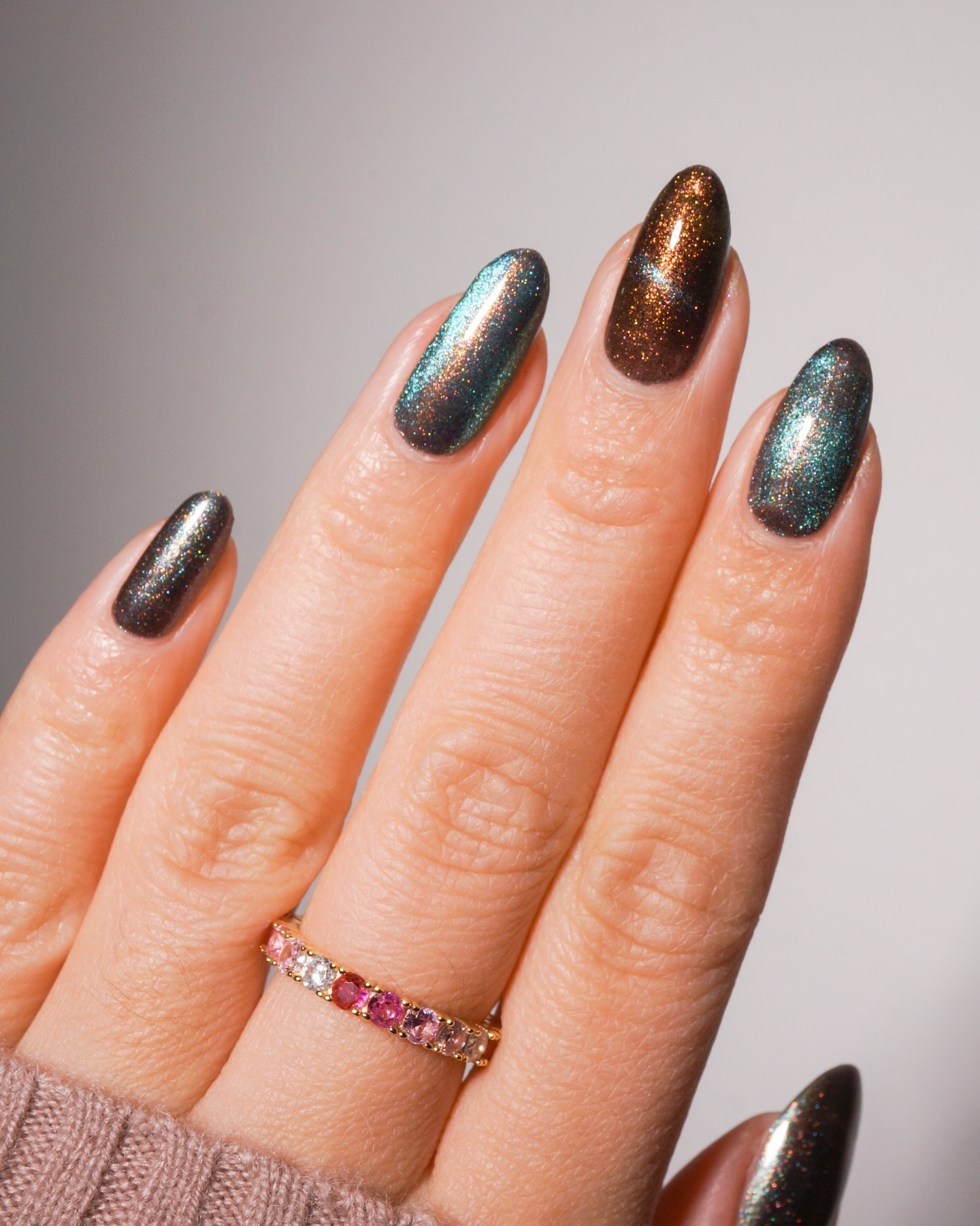 Unhas com esmalte cat eye, exibindo efeito metálico em tons esverdeados e dourados. Anel com pedras coloridas.